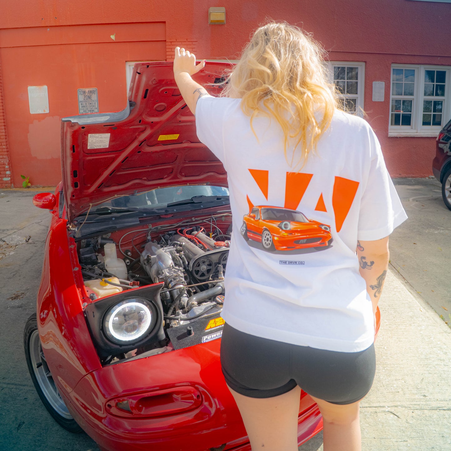 Person wearing a white t-shirt with red logo pointing at a red car with its hood open.