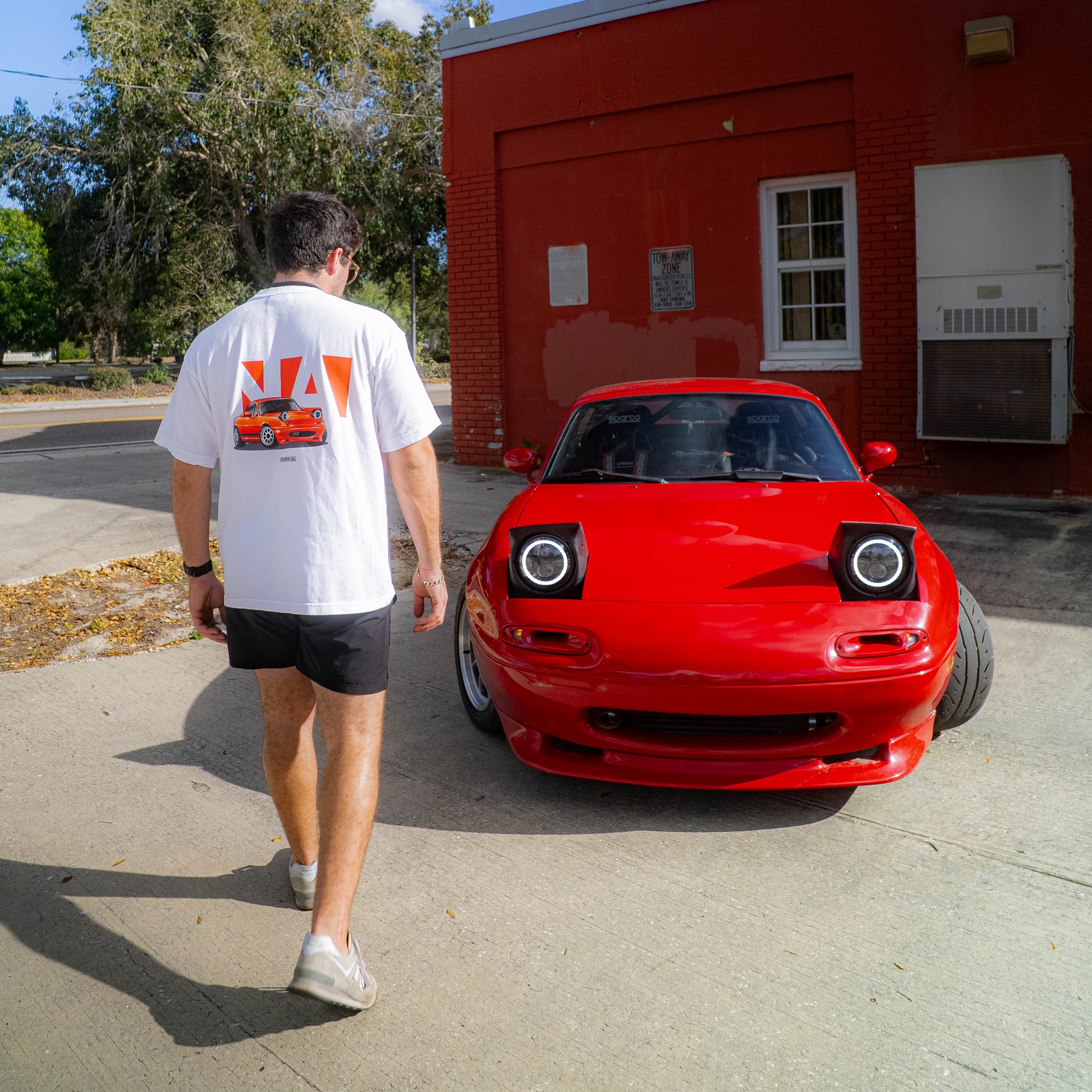 Person walking towards a red car on a sunny day with a building in the background