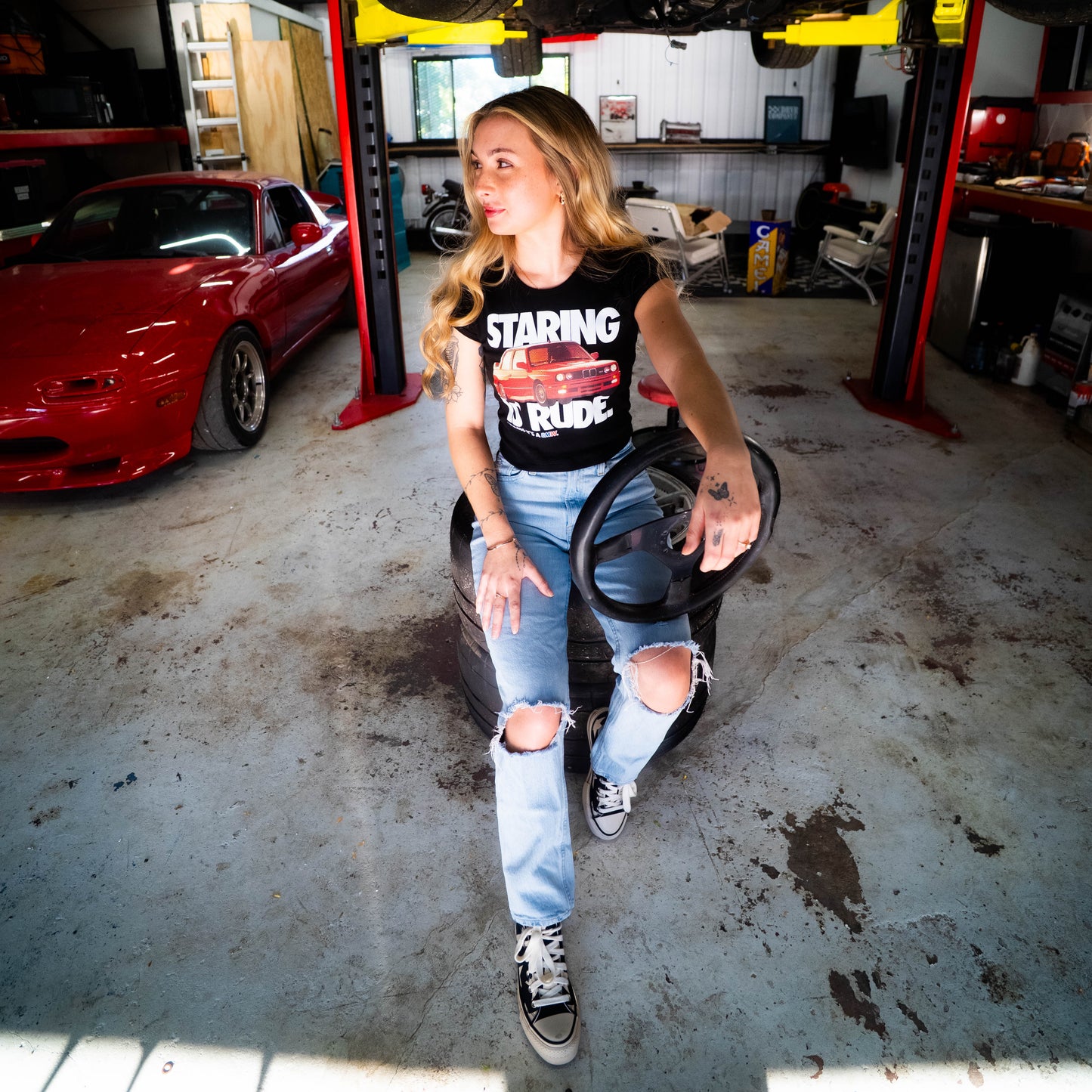 Person sitting on a car tire in a garage with a red car and tools in the background