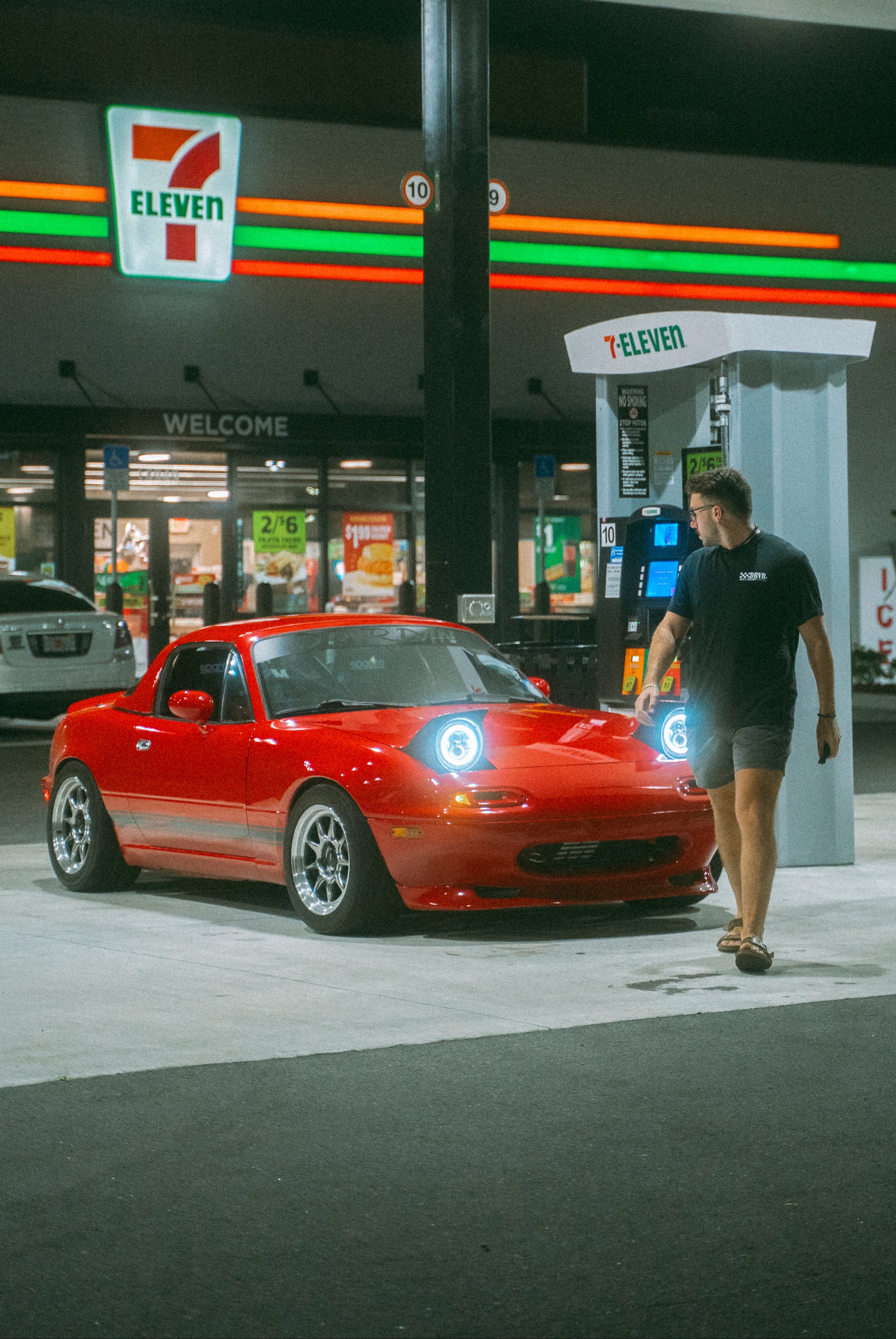 Red car parked at a 7-Eleven convenience store at night.
