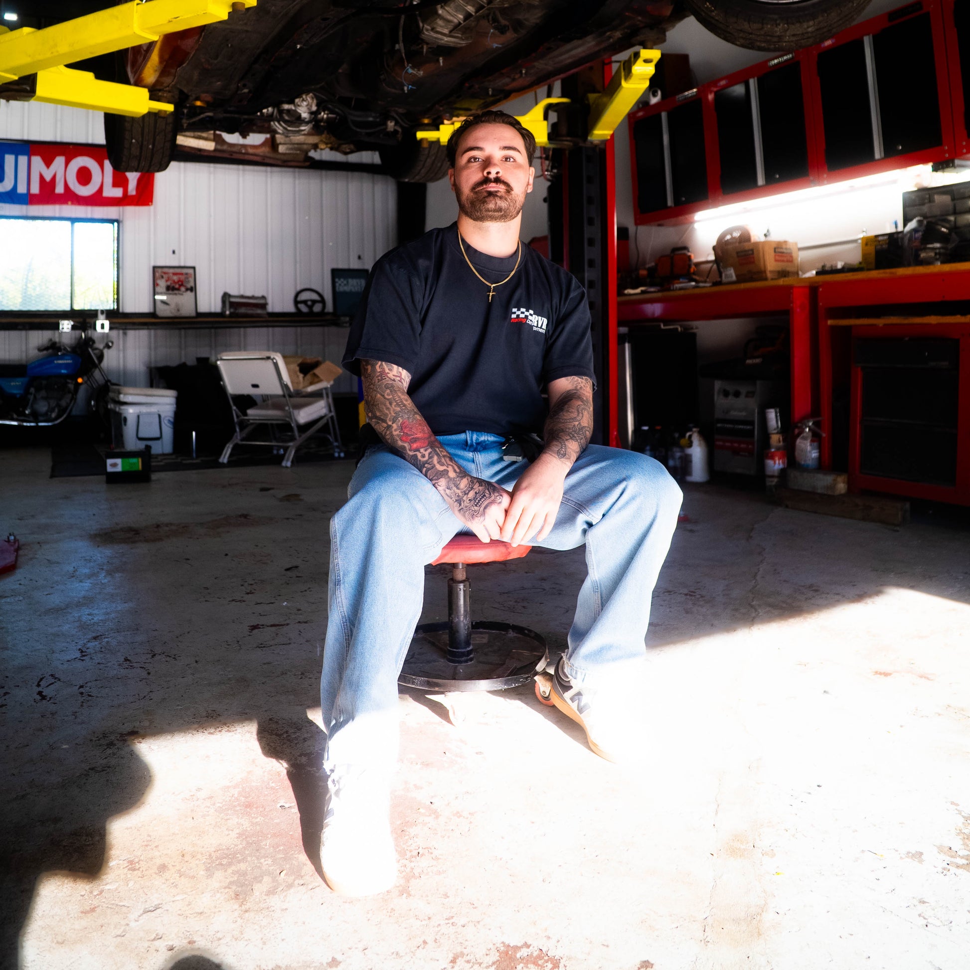 Mechanic sitting on a stool in a garage with a car lifted above.
