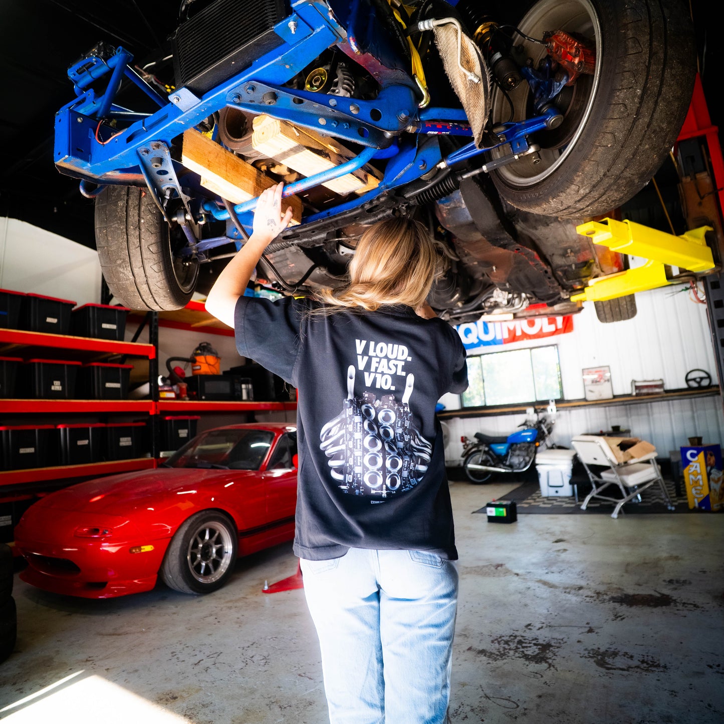 Person working on a car frame in a garage with various vehicles and equipment around.