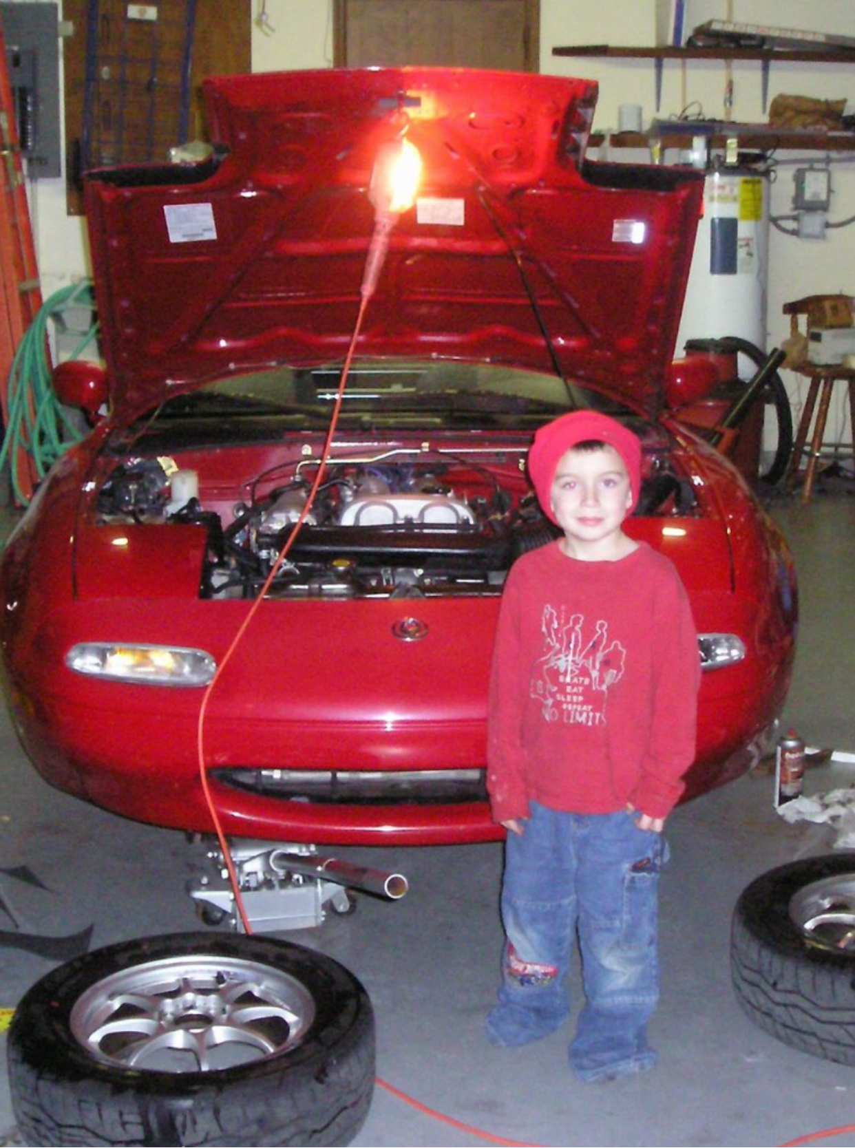 Child standing next to a red car with its hood open in a garage.