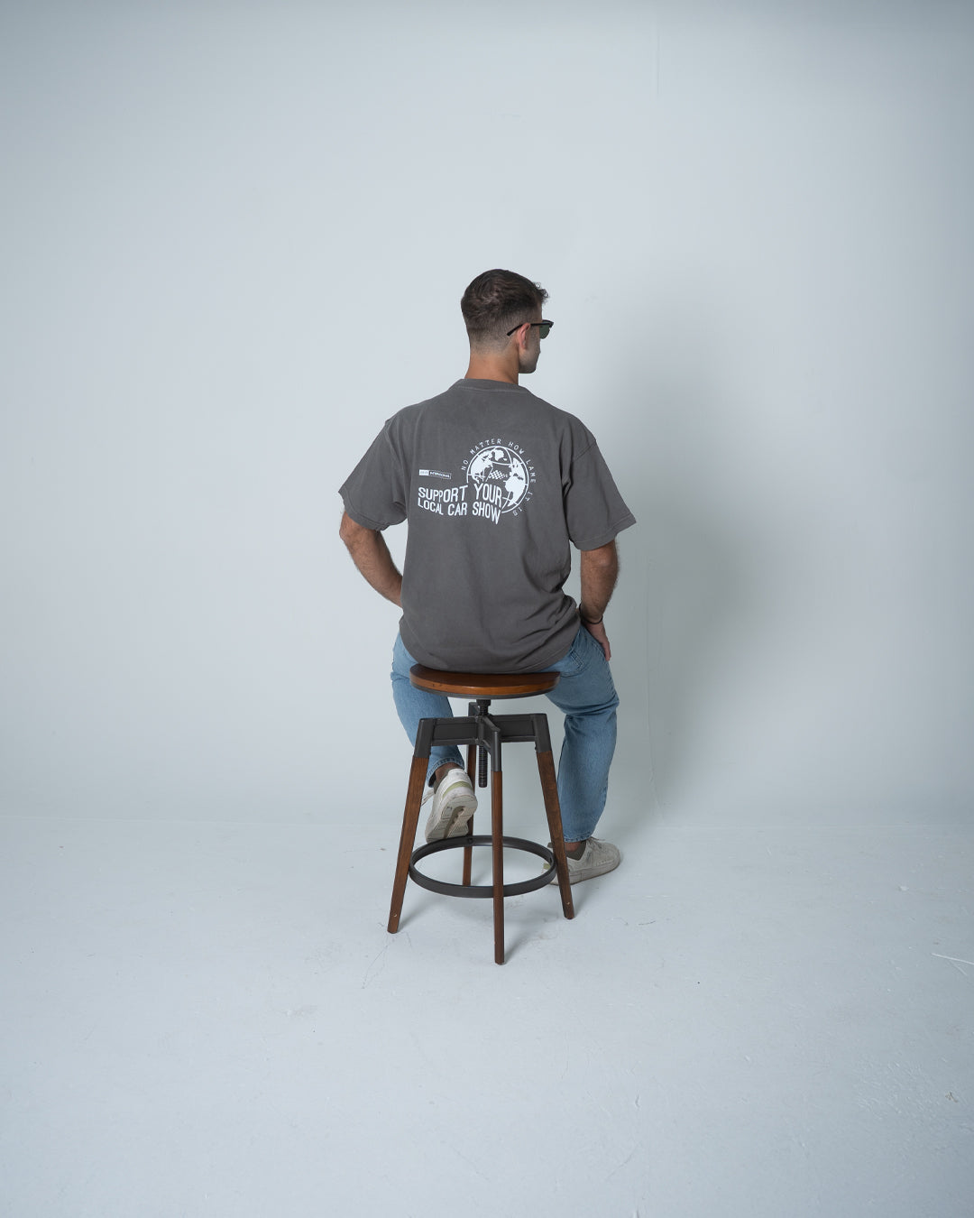 Man sitting on a stool wearing a gray t-shirt with a logo and text on the back.