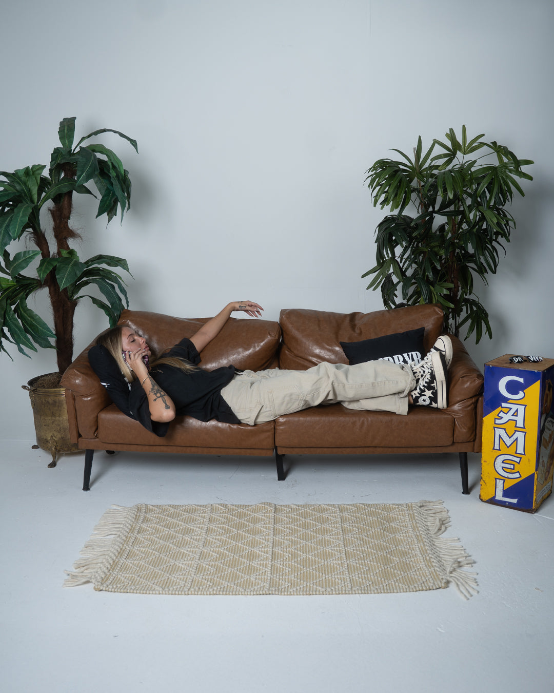 Person lying on a brown leather sofa with plants and a box in the background
